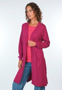 Fuchsia-Cardigan mit strukturiertem Strick, offener Front und langen Ärmeln. Verfügt über zwei frontale Taschen und ist über ein pink- und rotgestreiftes Oberteil geschichtet.