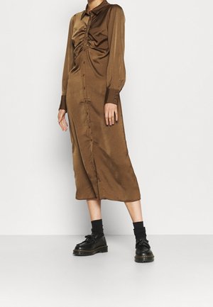 Vrouw die een bruin midi-jurk met lange mouwen en knopen draagt met geplooide details, zwarte sokken en zwarte plateauzolen, staande op een grijze vloer.