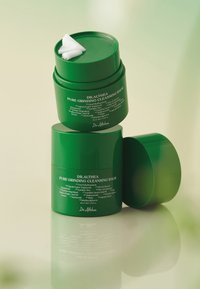 Zielone pojemniki na balsam, ułożone w stos, z lekko otwartą górną pokrywką, ukazującą biały produkt. Etykiety wyświetlają nazwę produktu i listę składników.