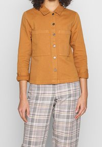 Persona vistiendo una camisa amarilla de botones con dos bolsillos en el pecho y mangas remangadas, combinada con pantalones blancos de cuadros con líneas negras y marrones.