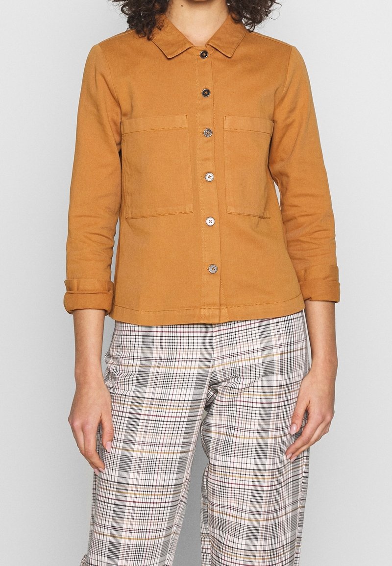 Persona vistiendo una camisa amarilla de botones con dos bolsillos en el pecho y mangas remangadas, combinada con pantalones blancos de cuadros con líneas negras y marrones.