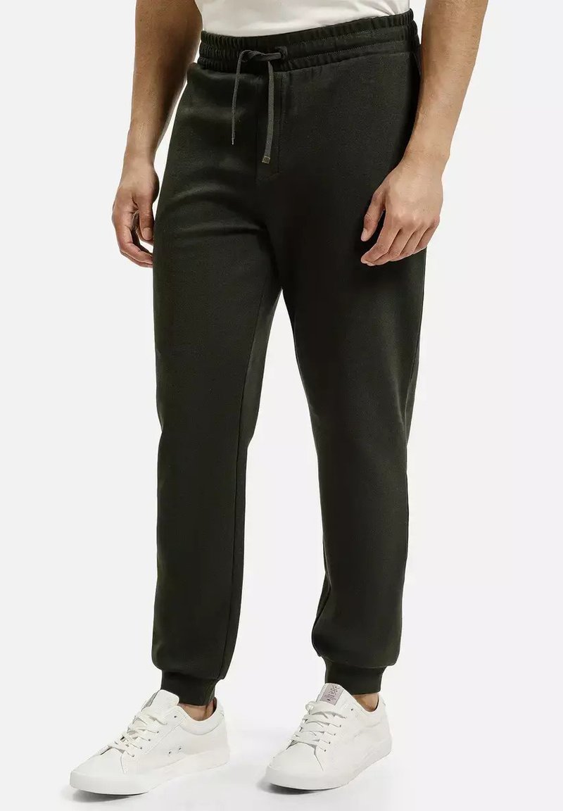 Ciemnozielone spodnie dresowe wykonane z miękkiego materiału, z elastycznym pasem ze sznurkiem oraz ściągaczami na końcach nogawek. Noszone z białymi trampkami.