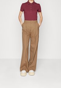 Personne debout avec les mains dans les poches, portant une chemise polo bordeaux, un pantalon beige taille haute et large, et des baskets blanches.