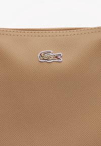 Tissu beige texturé avec logo crocodile cousu en marron clair et blanc avec détail rouge sur la bouche.