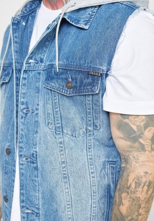 Denim-vesti, jossa on vaaleansinisiä ja haalistuneita kuvioita, kaksi rintataskua, napitus ja harmaa huppu, joka kurkistaa kauluksen alta.