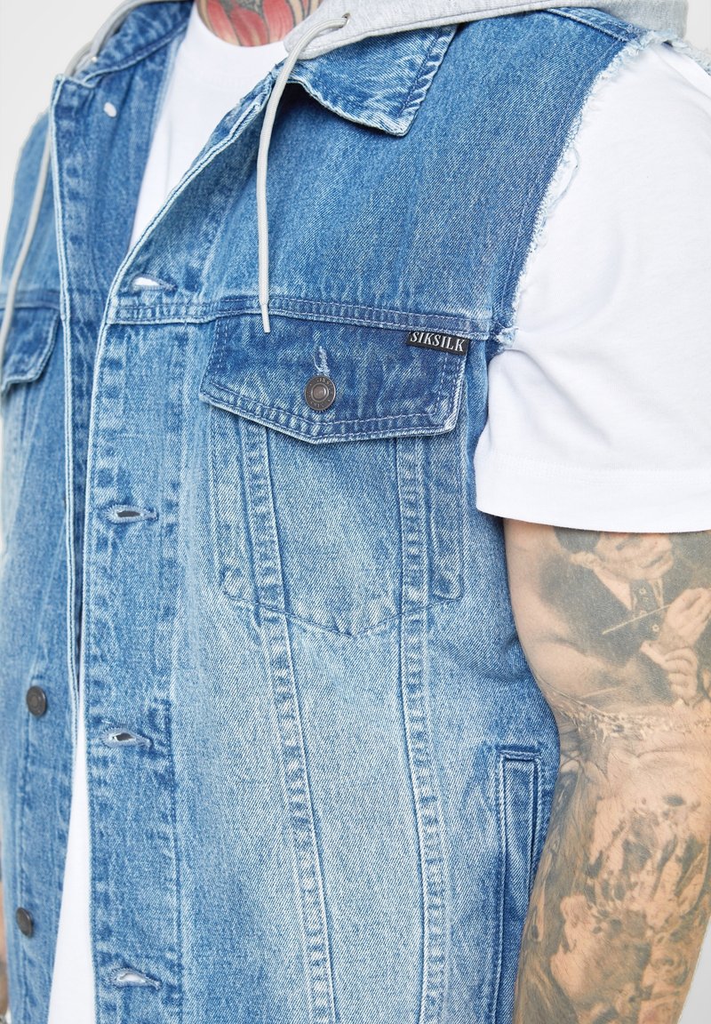 Jeansväst med ljusblå och bleknade texturer, två bröstfickor, knäppning med knappar och en grå huva som kikar fram under kragen.