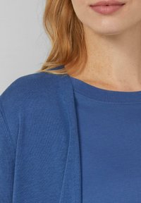 Niebieski sweter z dzianiny o fakturowanym splocie, połączony z gładką niebieską koszulką. Posiada luźny krój i szeroki dekolt.