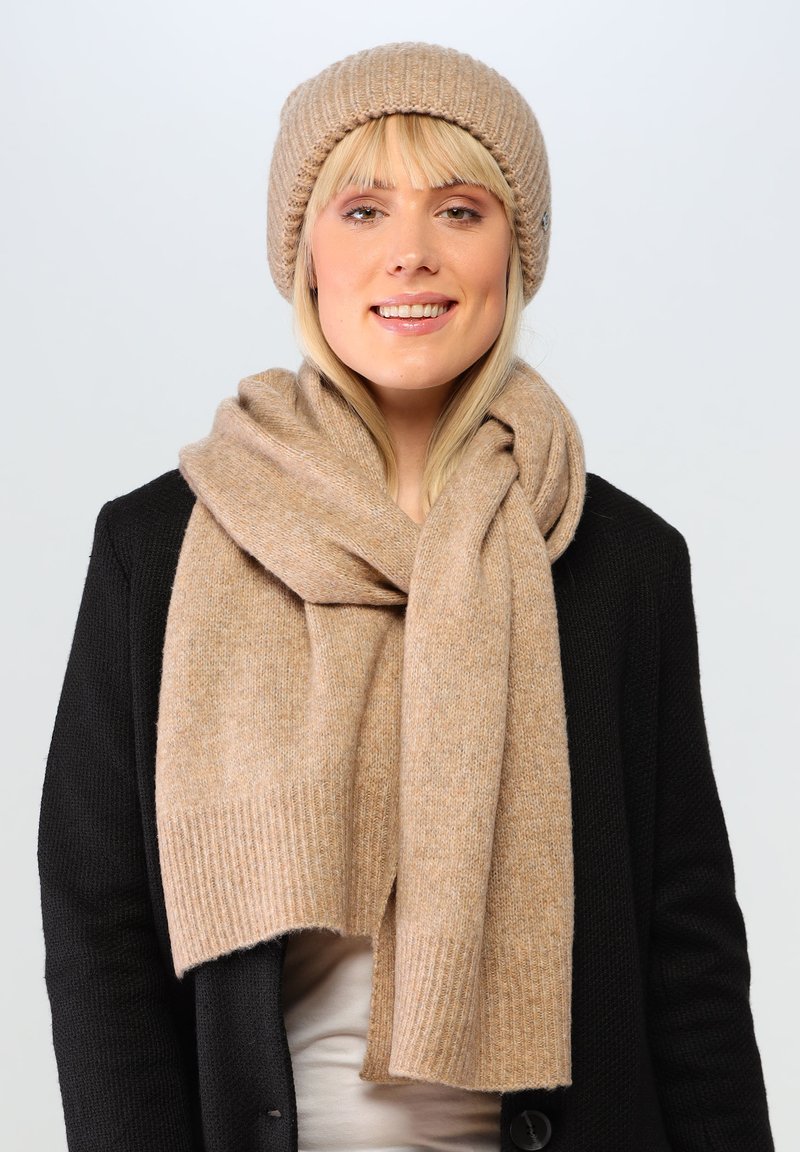 Beanie beige in maglia e sciarpa coordinata realizzati in morbida lana, con texture a costine. Indossati sopra un cardigan nero su uno sfondo chiaro.