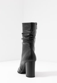 Černá kožená kotníková bota s uvolněným designem, zapínáním na zip, s kulatou špičkou a robustním podpatkem. Hladký povrch s jemnými švy.