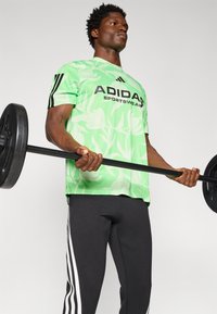 Žalias gėlėmis raštuotas Adidas sportinis džemperis, juodos sportinės kelnės su baltomis juostelėmis, kelia barbell'į, raumeninga kūno sudėtis, neutralus fonas.