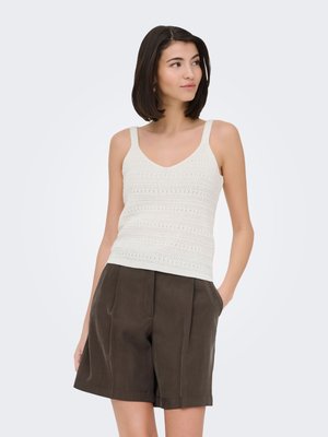 Vrouw met kort donker haar die een witte gebreide tanktop en losse donkerbruine shorts met hoge taille draagt, staand met één hand in de zak.