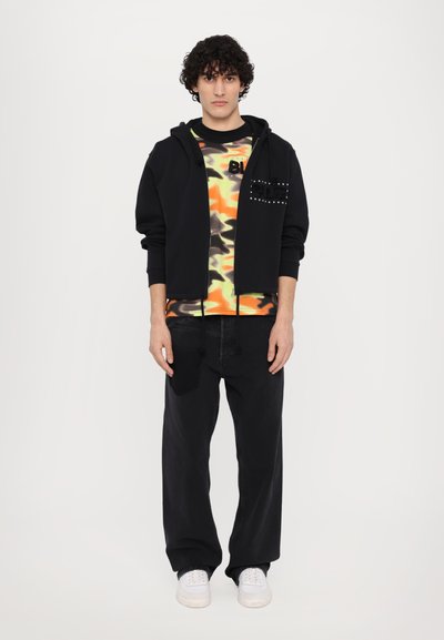 Haut à capuche zippé noir avec des détails cloutés, t-shirt camouflage orange et vert en dessous, jean noir à jambe large et baskets blanches.
