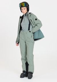 Helle grüne Schneejacke mit Reißverschlusstaschen, kombiniert mit passenden Schneehosen mit verstellbaren Trägern und schwarzen Skischuhen.