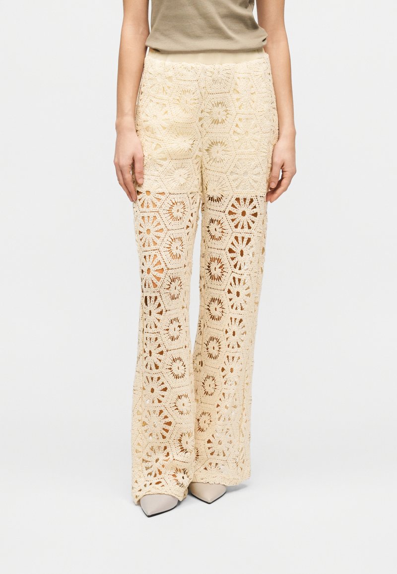 Εκρού πλεκτό παντελόνι φαρδιάς γραμμής σε δαντέλα, συνδυασμένο με μπεζ μπλούζα και μπεζ παπούτσια με μυτερά δάχτυλα, εμφανίζεται από τη μέση μέχρι τα πόδια.