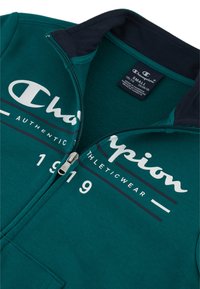 Giacca a zip verde acqua con colletto blu navy; presenta il logo bianco "Champion", il testo "AUTHENTIC ATHLETICWEAR" e l'anno "1919" in colori contrastanti.
