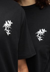 Μαύρα κοντομάνικα μπλουζάκια με λευκό ανθικό σχέδιο στο αριστερό στήθος. Το ύφασμα φαίνεται λείο με κανονική εφαρμογή.