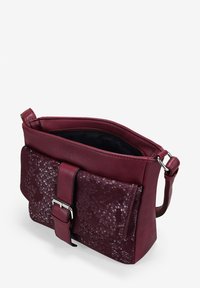 Bolso de hombro en cuero rojo oscuro con solapa frontal texturizada y hebilla plateada, parcialmente abierto mostrando el forro interior negro.