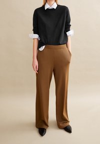 Pull noir à manches retroussées superposé à une chemise blanche, associé à un pantalon large marron. Présente des coutures discrètes et des poches latérales.