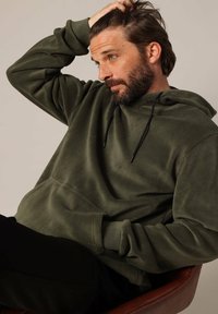 Olijfgroene fleece hoodie met een pouchzak aan de voorkant, verstelbare zwarte trekkoorden en ribgebreide manchetten. Zachte textuur en een relaxte pasvorm.