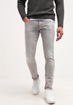 Homme portant un jean slim gris clair, un pull en maille gris foncé, des baskets blanches, marchant avec une main dans la poche sur fond uni.