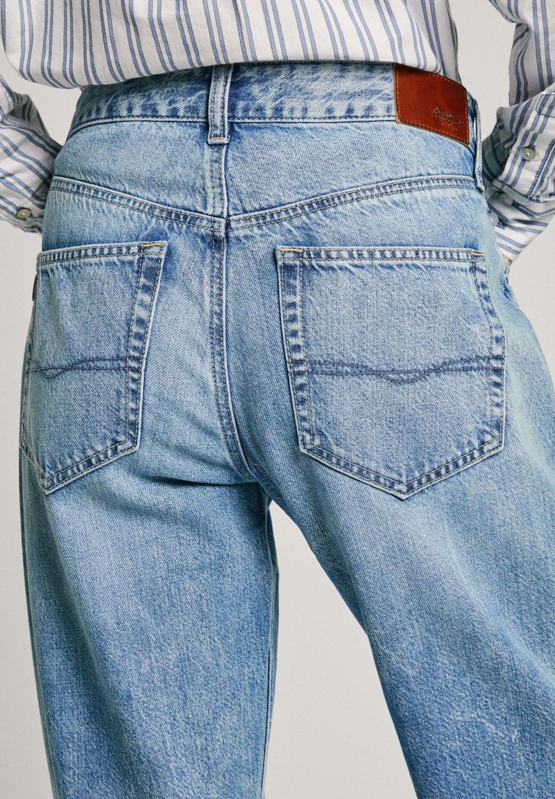 Jeans in denim blu chiaro con vita alta, due tasche posteriori e una patch in pelle marrone sulla cintura. Il tessuto ha una texture sbiadita.