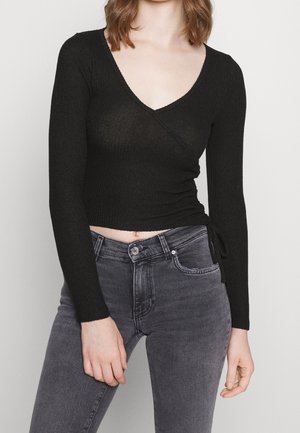 Kvinde iført en sort langærmet omslagstop med V-hals kombineret med højtaljede falmede sorte jeans, stående mod en ensfarvet baggrund.