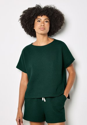 Frau mit lockigem Haar, die ein dunkelgrünes, strukturiertes Kurzarm-Top und passende Shorts trägt, steht mit einer Hand in der Tasche vor einem einfarbigen Hintergrund.