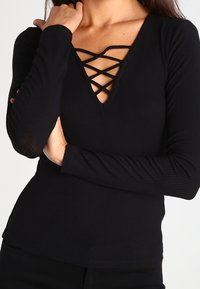 Černý dlouhý rukáv top s ribbovanou texturou, hlubokým V-výstřihem s kříženými popruhy, upnutým střihem a bezšvým lemem.