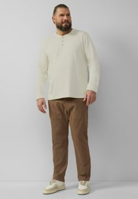 Crème lange mouwen henley-shirt met drie knopen, gecombineerd met bruine cargobroek en crème sneakers, tegen een lichtgrijze achtergrond.