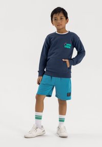 Sweatshirt bleu marine avec texte vert, équipé d'une poche avant ; associé à un short turquoise et des chaussures blanches avec des chaussettes à rayures vertes.