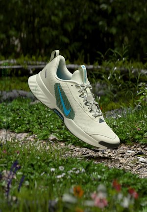 Lichtbeige Nike Trail hardloopschoen met blauw logo, zwevend boven een grind- en grasachtig pad omgeven door groene planten en wilde bloemen.
