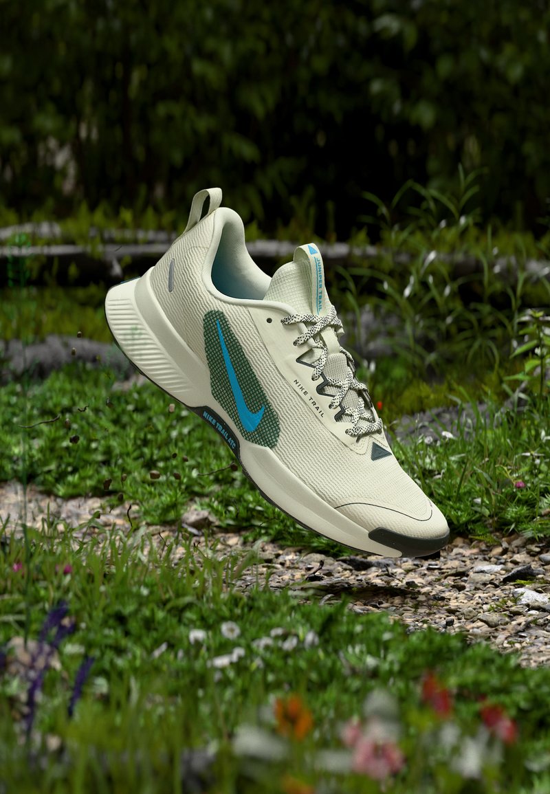 Ανοιχτό μπεζ παπούτσι για τρέξιμο Nike Trail με μπλε λογότυπο, αιωρείται πάνω από ένα μονοπάτι με χαλίκι και γρασίδι, περιτριγυρισμένο από πράσινα φυτά και άγρια λουλούδια.