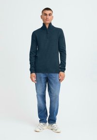 Dunkelblauer Strickpullover mit hohem Kragen, schmalen Ärmeln und geripptem Saum, kombiniert mit blauen Jeans und weißen Sneakers.
