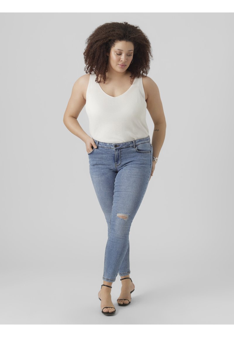 White ribbed tank top paired with light blue skinny jeans featuring a knee rip. Model wears black ankle strap sandals, with a neutral background.