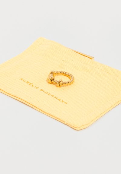 Χρυσό δαχτυλίδι σε στυλ στριμμένης κορδέλας με δύο λεπτομέρειες κόμπων, πάνω σε κίτρινη υφασμάτινη θήκη με την ετικέτα Aurélie Bidermann.