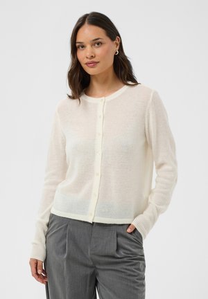 Femme aux cheveux foncés porte un cardigan blanc boutonné et un pantalon plissé gris, debout avec une main dans la poche devant un fond uni.