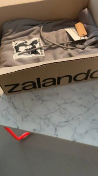 Sweatshirt cinza dobrado dentro de uma caixa castanha da Zalando, apresentando uma impressão gráfica de um rosto e a palavra "FEARLESS". Materiais são macios e descontraídos.