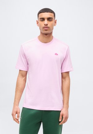 Mees seisab näoga ettepoole, kandes heleroosat lühikeste varrukatega särk ja tumerohelisi pükse ühetaolise heleda tausta ees.