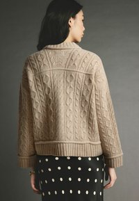 Kabelstrick-Cardigan in hellbeige, mit geripptem Saum und Bündchen. Getragen über einem schwarzen Rock mit weißen Punkten.