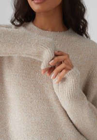 Maglione beige, con una texture morbida e pelosa, e polsini a costine. Dettagli scintillanti sono visibili su tutta la superficie del tessuto.