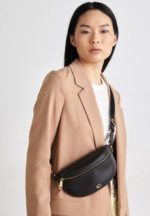 Kvinde iført beige blazer og hvid top med sort læder crossbody taske med guldfarvet lynlås og spænder.