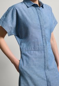Abito in chambray con colletto a camicia, maniche corte e vita sagomata. Presenta una chiusura frontale con bottoni e una texture liscia e leggera.