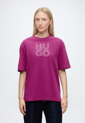 Blonde Frau trägt ein locker sitzendes lila T-Shirt mit "Hugo"-Logo und dunkle Hose, steht vor schlicht weißem Hintergrund.