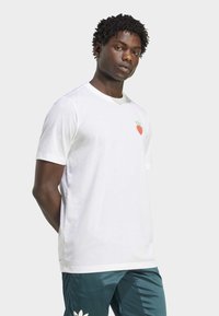 Camiseta blanca de manga corta con un pequeño gráfico de fresa roja en el lado izquierdo del pecho. La tela parece suave y ligera.