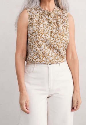 Ærmeløs brun bluse med hvidt blomsterprint og knapper, båret med højtaljede hvide bukser på en person med langt gråt hår.