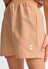 Helle beige Shorts aus glattem Stoff, mit einem elastischen Bund, zwei seitlichen Taschen und einem weißen Chevron-Logo an der unteren Seite.