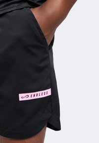 Homme avec la main dans la poche portant un short noir avec le logo rose "ENDLESS" sur la cuisse gauche, fond uni.