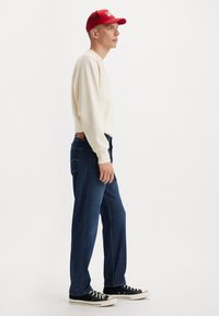 Camisa blanca de manga larga, jeans azul oscuro y zapatillas negras. Gorra roja con texto blanco. Vista lateral, el modelo está de pie sobre un fondo liso.