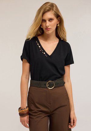 Blonde vrouw in een zwart V-hals t-shirt met edelsteen details, bruine broek met een brede riem, gouden hoepel oorbellen en houten armbanden om haar linker pols.
