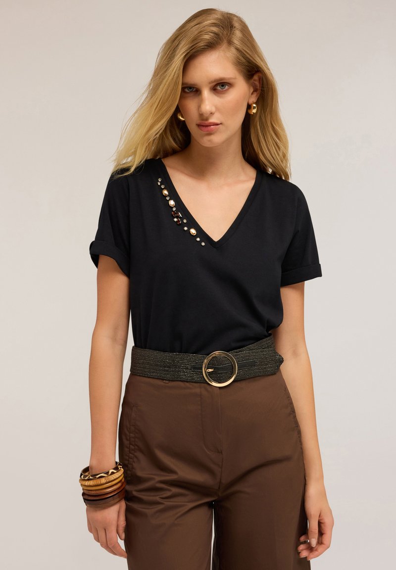 Mujer rubia con camiseta negra de escote en V con detalles de piedras preciosas, pantalones marrones con cinturón ancho, pendientes de aro dorados y pulseras de madera en la muñeca izquierda.